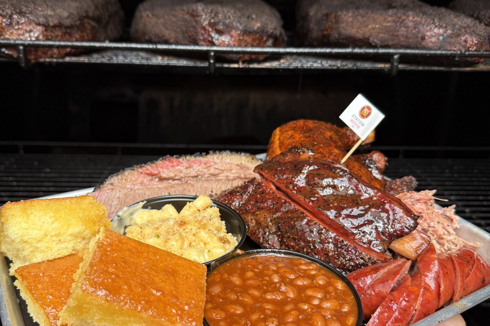 A Station House BBQ tray of sliced brisket, ribs, sausage, and chicken with Smoked Gouda Mac & Cheese, Cowboy Baked Beans, and cornbread, held in front of the smoker